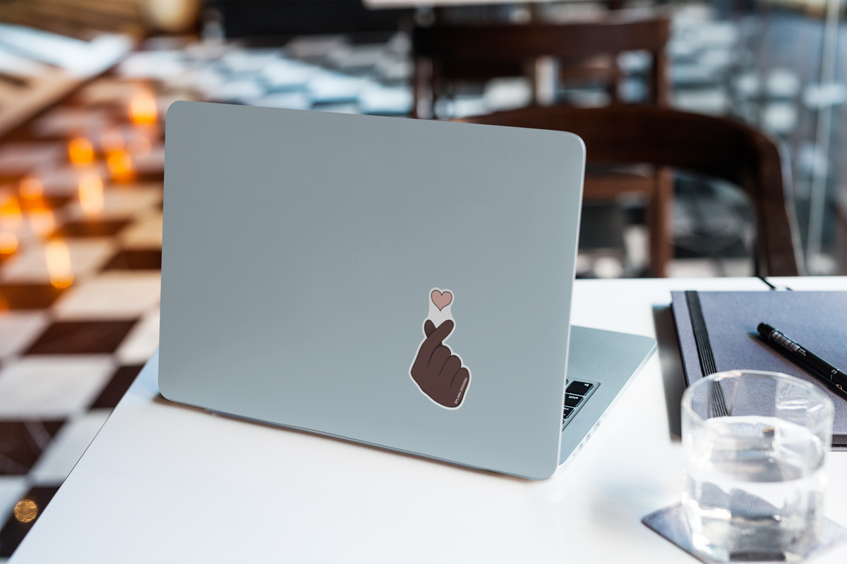 Autocollant cœur de doigt de la marque Prodigi appliqué sur une couverture d'ordinateur portable, posé à côté d'un verre d'eau.